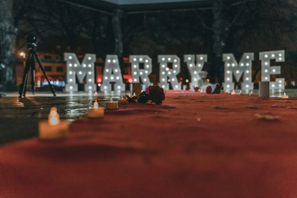 A romantic setup for a marriage proposal with lighted "Marry Me" sign and candles at night.